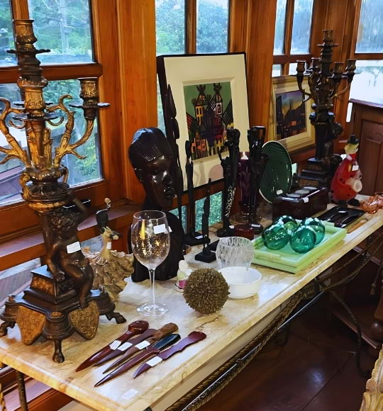 Mesa lateral com candelabros antigos, esculturas, quadros, taças e objetos de decoração raros, destacados em ambiente com luz natural na Coar.